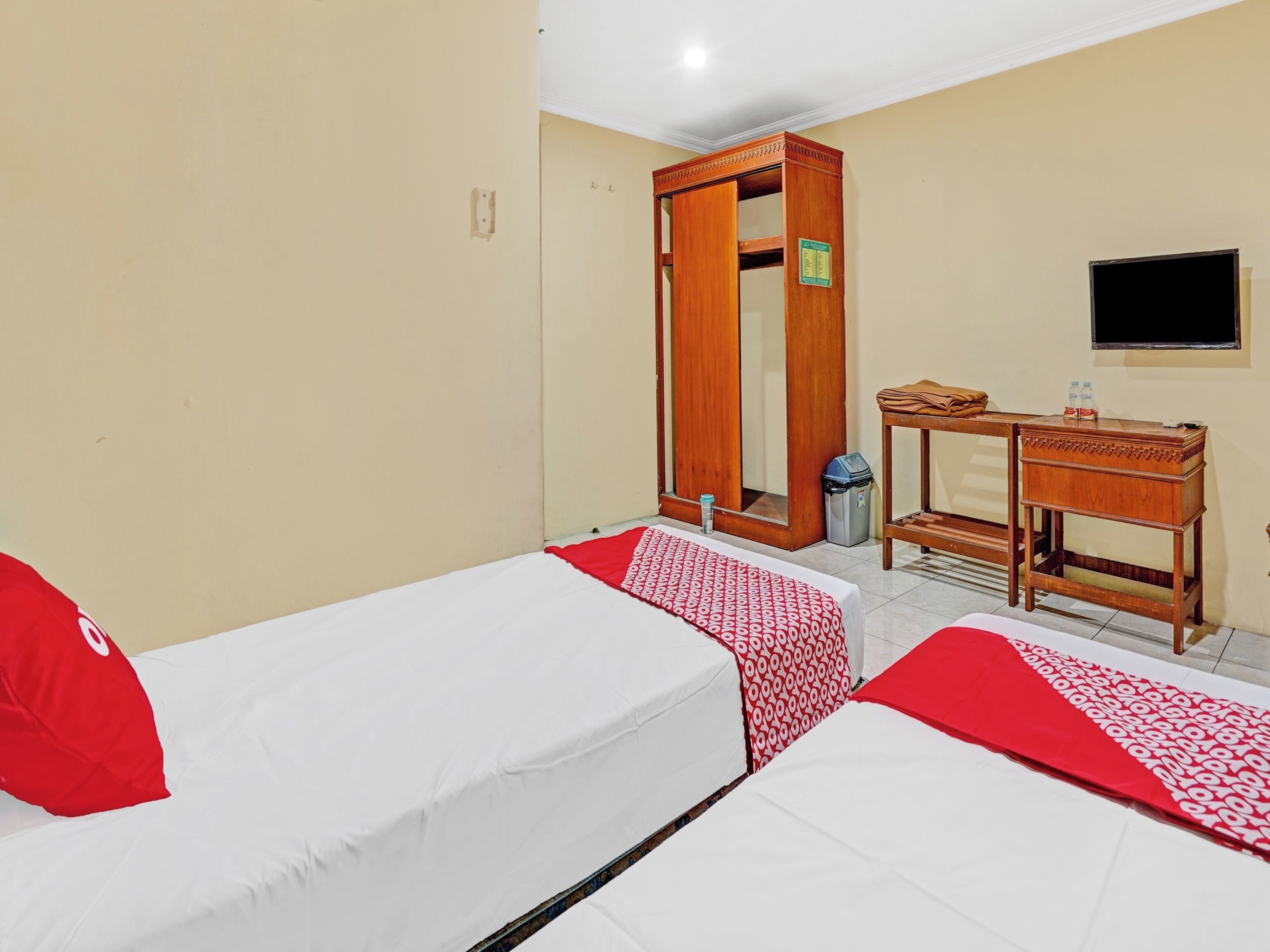 A hotel room with two beds, a wooden wardrobe, a desk, a trash bin, and a television on the wall.