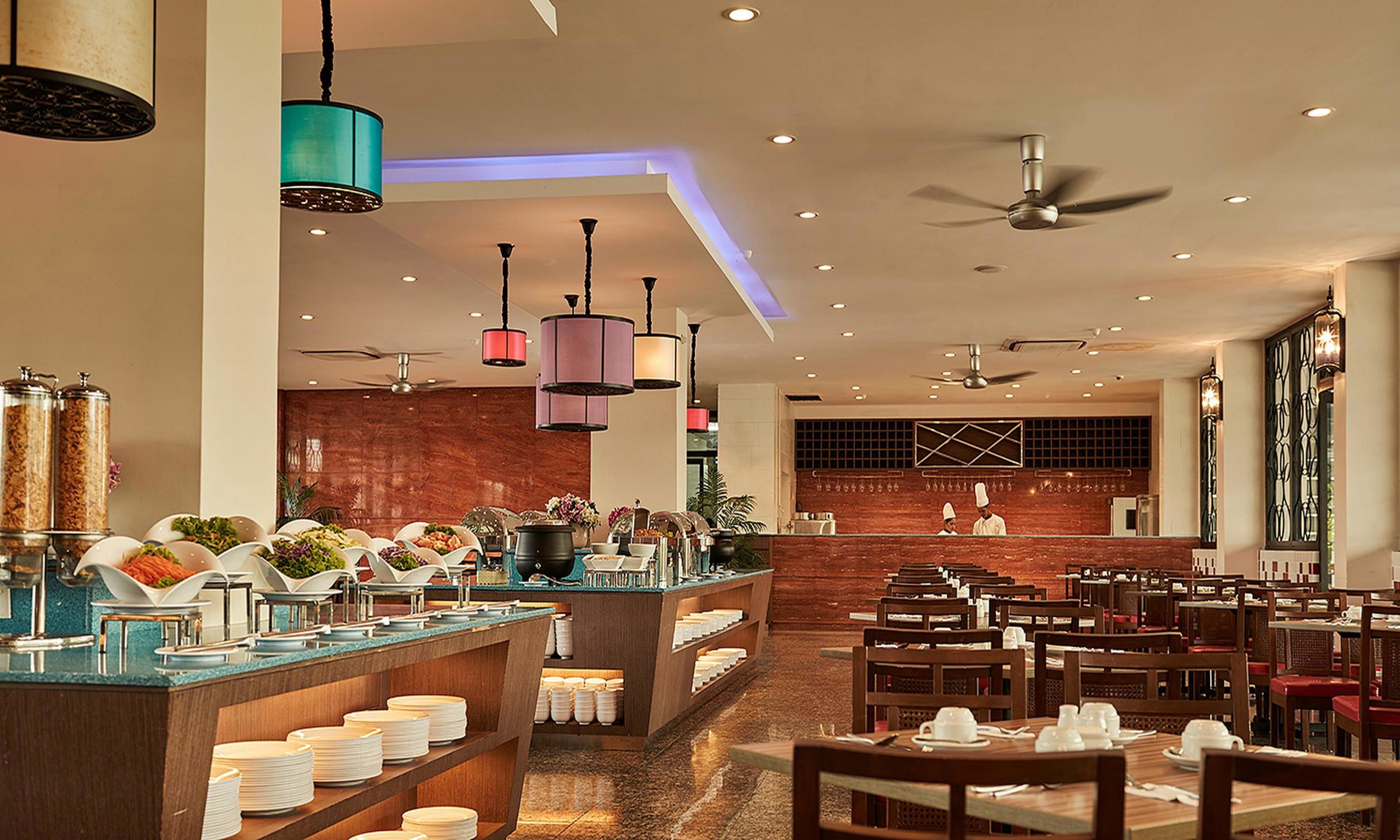 A hotel restaurant with a buffet area, dining tables, and a kitchen in the background.