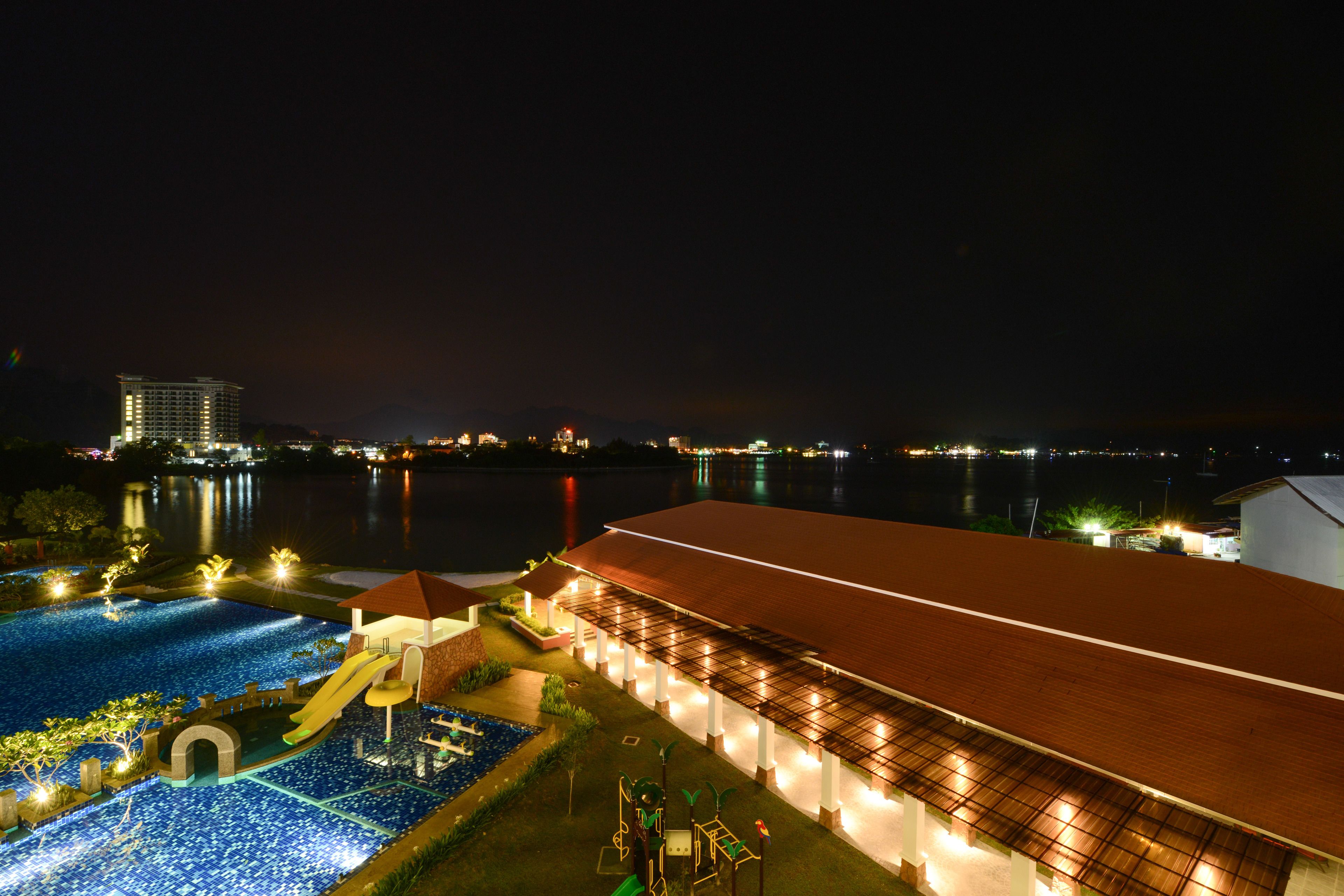 A nighttime view of a hotel with a swimming pool, a covered area, and a view of the water and distant buildings.