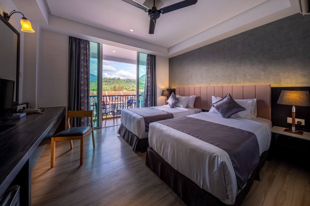 Une chambre d’hôtel avec deux lits, un bureau, une chaise, un ventilateur de plafond et un balcon offrant une vue sur les montagnes.