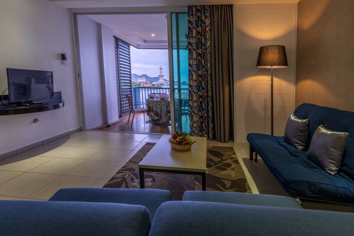 Un salon moderne avec un canapé, un téléviseur, une table sur laquelle se trouve un panier de fruits, et une vue sur un balcon doté d’une balustrade et sur une tour située au loin.