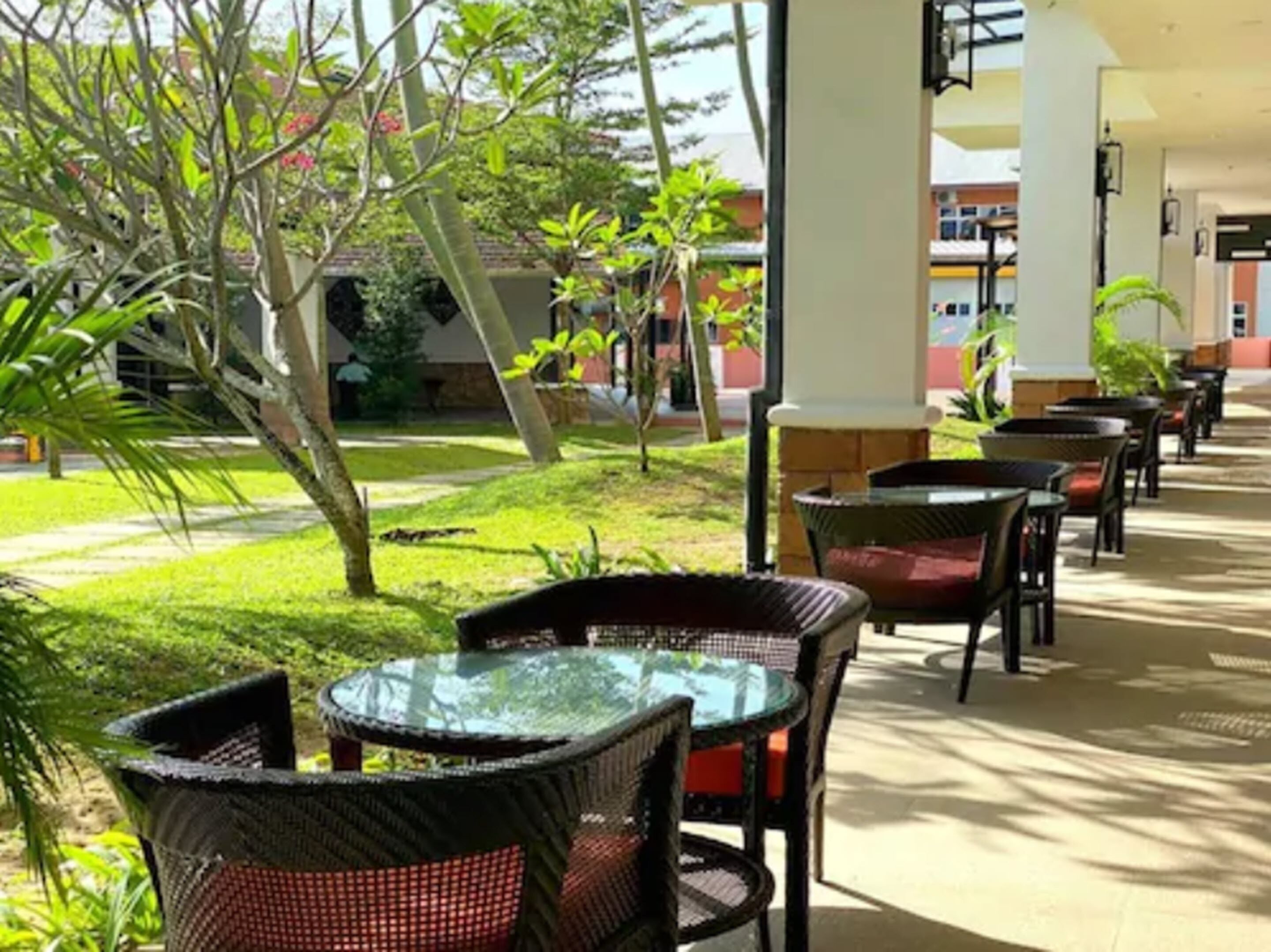 A covered outdoor seating area with wicker chairs and tables, surrounded by greenery and buildings in the background.