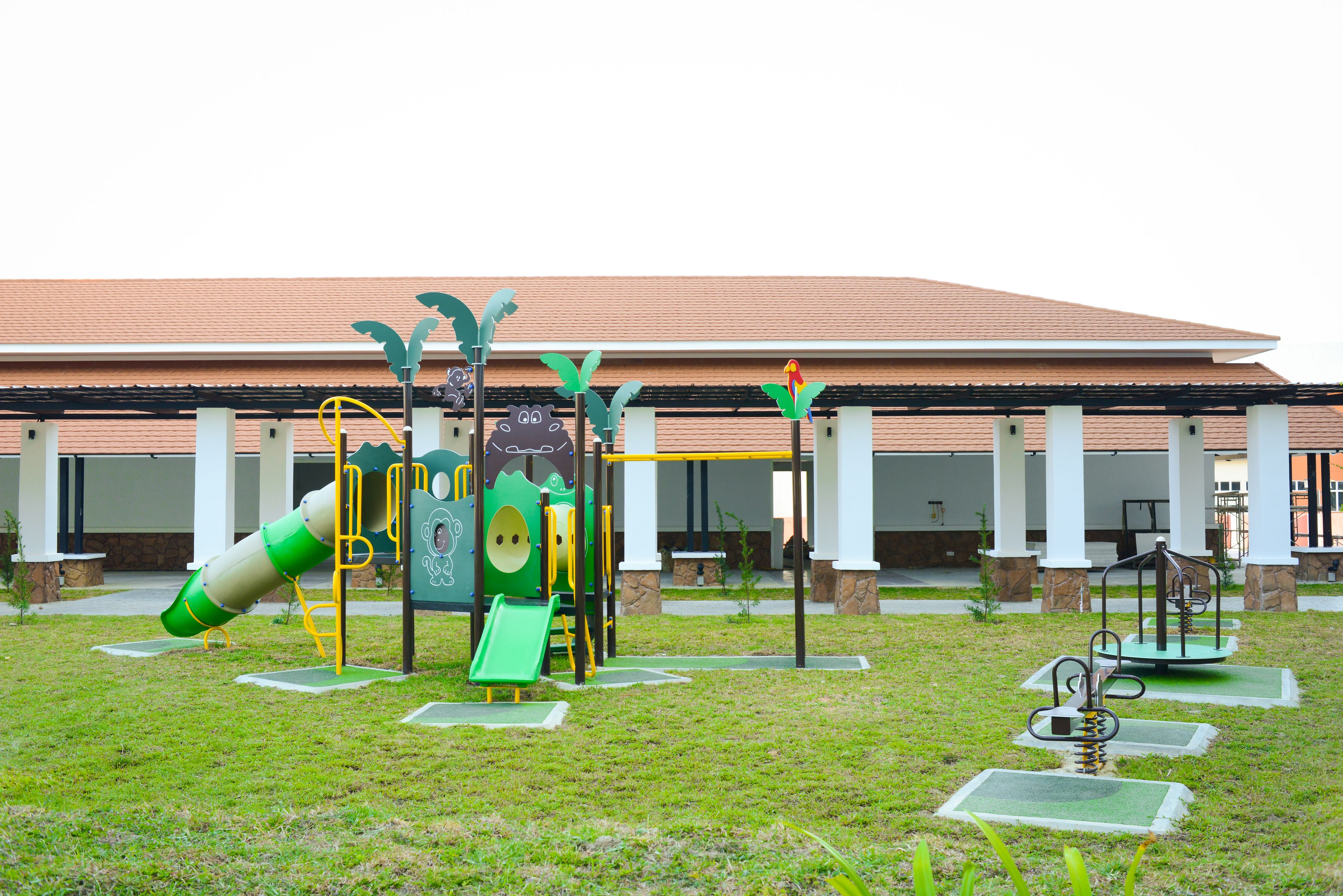 Un parque infantil con diversas estructuras de juego, incluyendo un tobogán y zonas de escalada, frente a un edificio con tejado de baldosas y columnas.