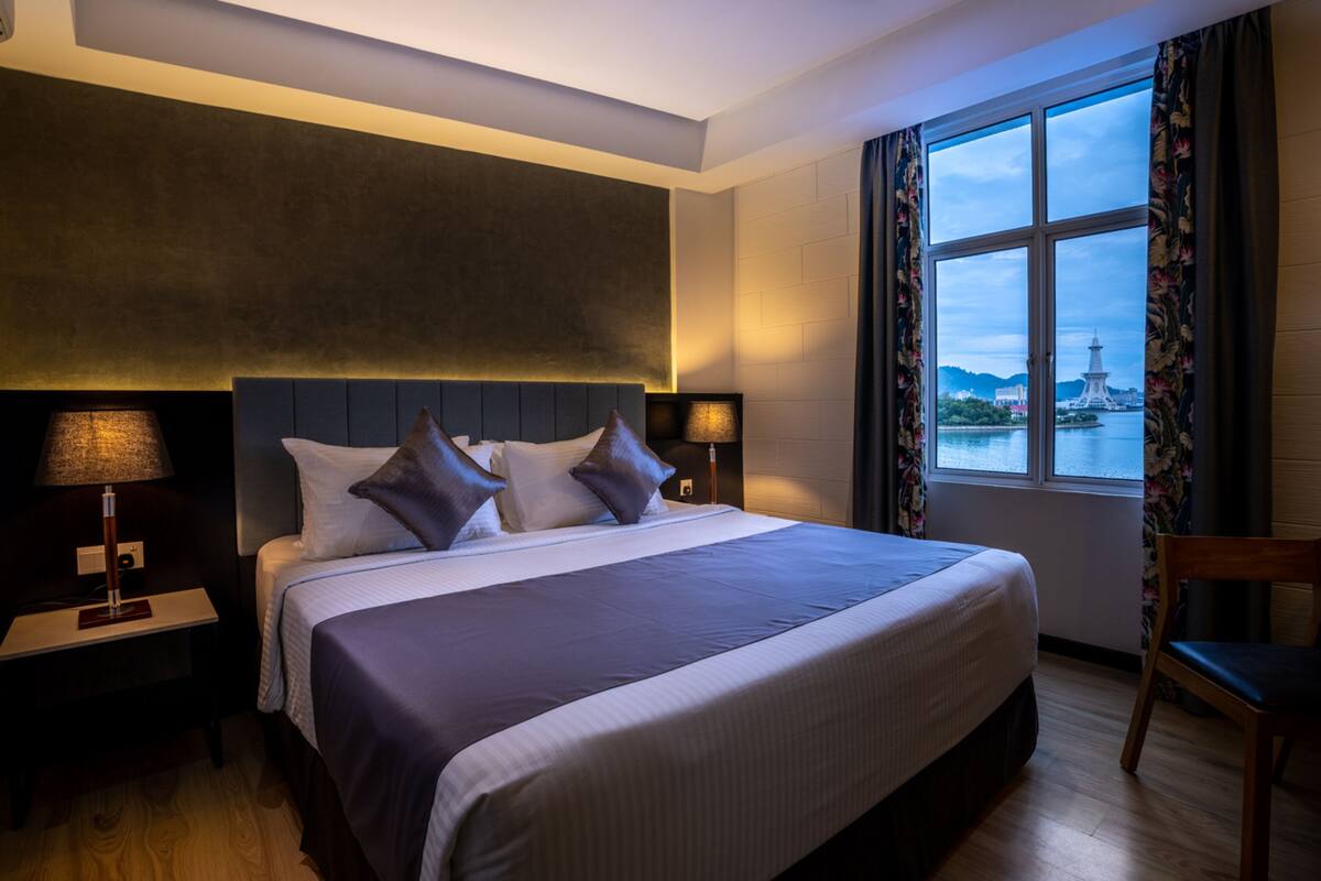 Une chambre d’hôtel moderne avec un grand lit, une table de chevet avec une lampe, une chaise et une fenêtre donnant sur une tour située au loin.