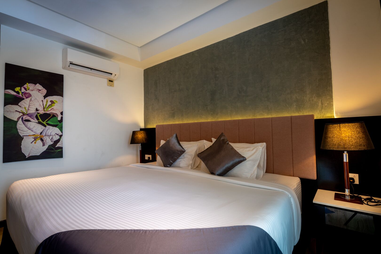 Et hotelværelse med en stor seng, to sengeborde med lamper, en vægmonteret aircondition og et stort abstrakt maleri over sengen.