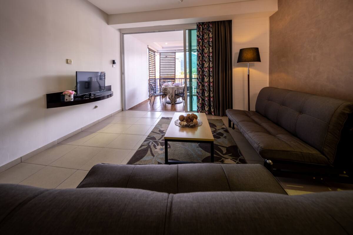 Un salon moderne avec un canapé, une table basse agrémentée d’objets décoratifs, un téléviseur à écran plat posé sur une étagère, et une vue sur un balcon extérieur.