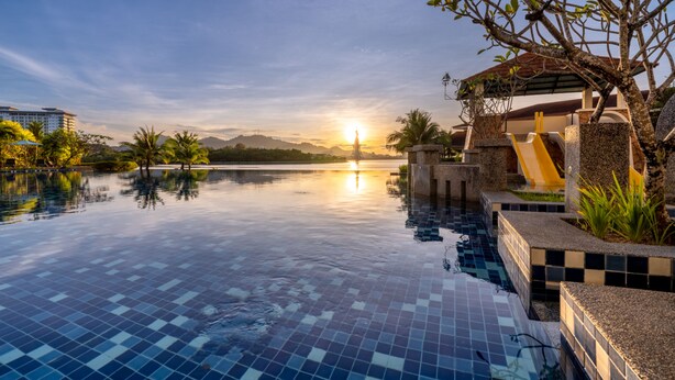 Une piscine à débordement paisible, offrant une vue dégagée sur le coucher de soleil au-dessus d’une chaîne de montagnes, entourée d’une végétation luxuriante et d’une structure architecturale moderne.