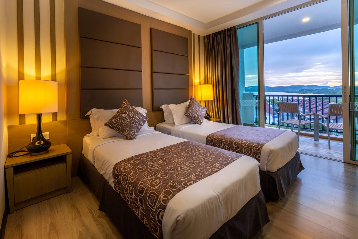 Une chambre d’hôtel avec deux lits, une table de chevet, une lampe, une vue sur le balcon et une grande fenêtre.