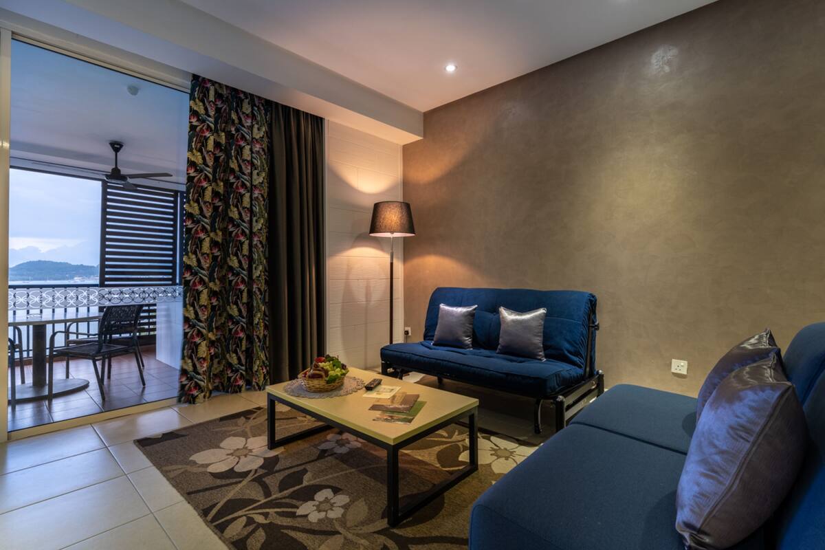 Un salon moderne avec un canapé bleu, une table basse et un lampadaire. Il y a un balcon avec vue sur la mer et les montagnes.