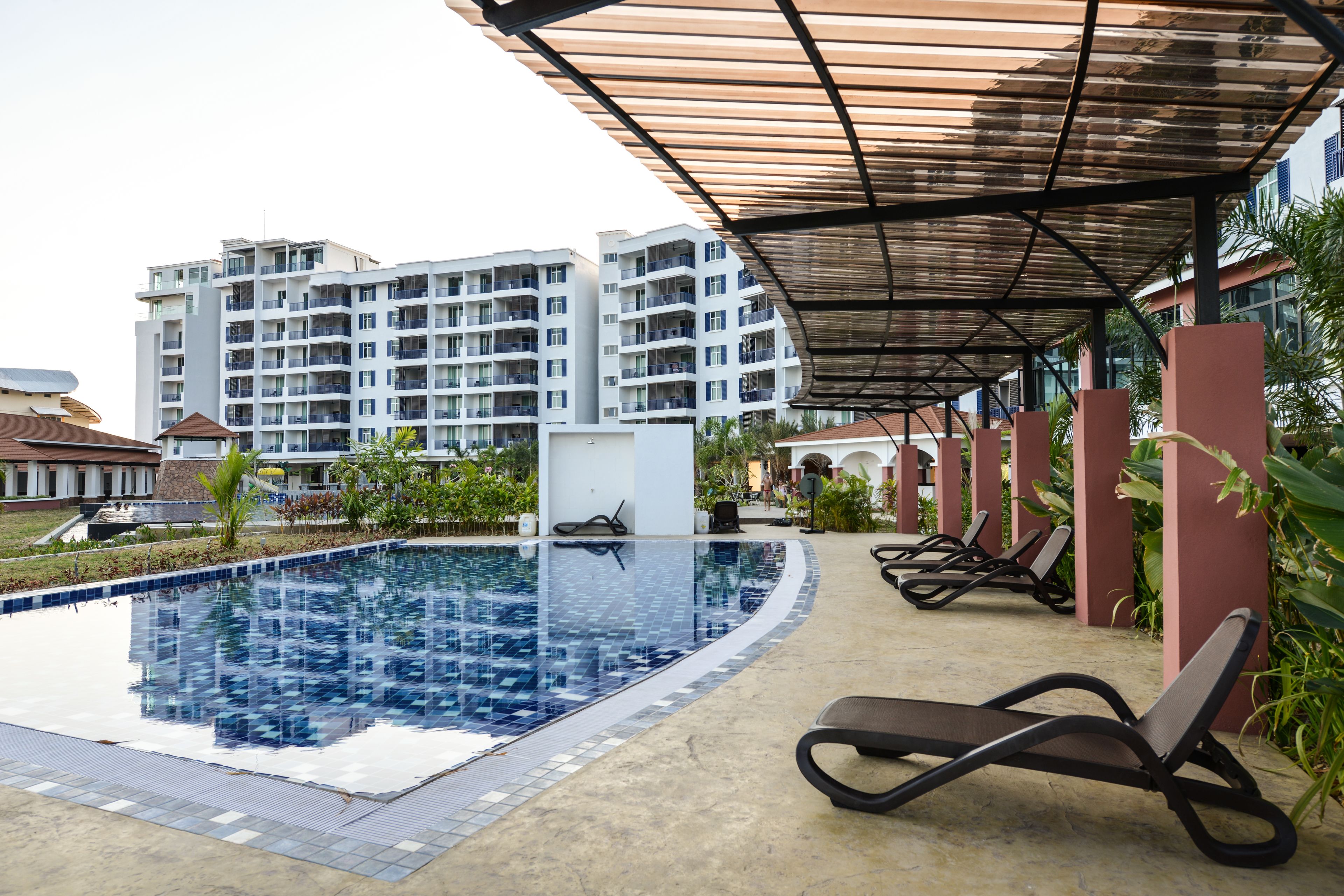 A swimming pool with lounge chairs and a covered area, surrounded by buildings and greenery.