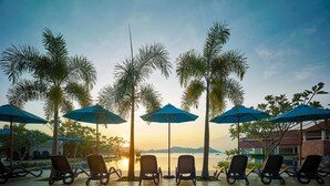 Un espace aménagé au bord de la piscine, avec des chaises longues et des parasols, offrant une vue sur une étendue d’eau et des collines lointaines au coucher du soleil.