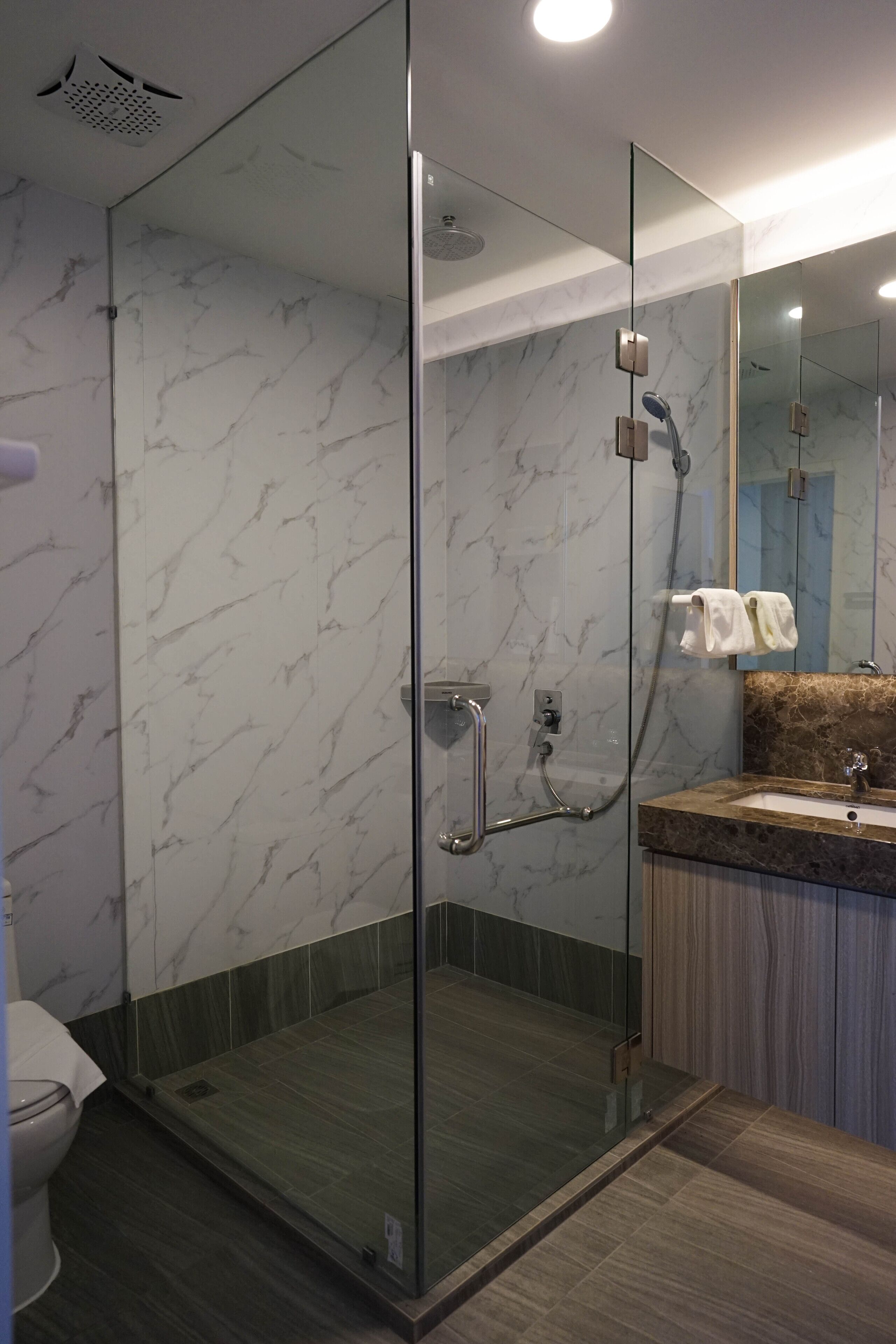 Un baño moderno con ducha envuelta en vidrio, paredes de mármol y tocador de madera.