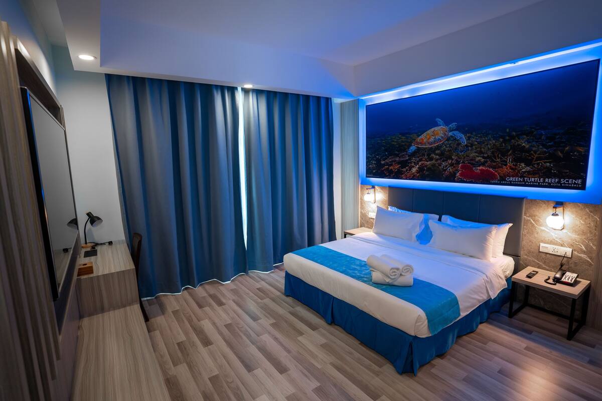 A modern hotel room with a large bed, a flat-screen TV displaying a marine scene, a desk with a lamp, and a nightstand with a phone and lamp.