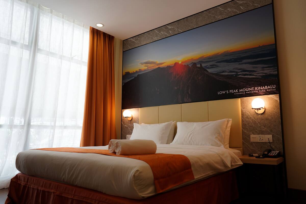 A hotel room with a large bed, orange curtains, a wall-mounted light, and a poster of Mount Kinabalu.