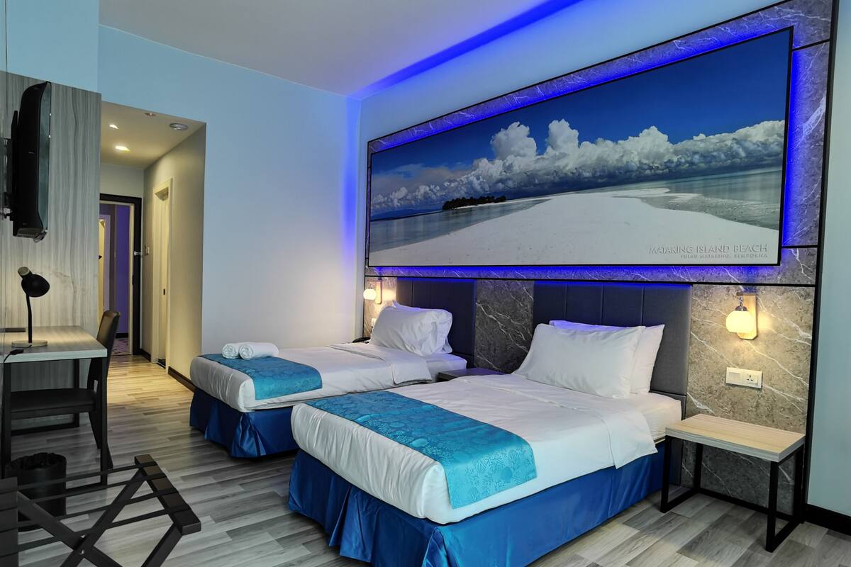 A hotel room with two beds, a desk, and a large TV screen displaying a beach scene.