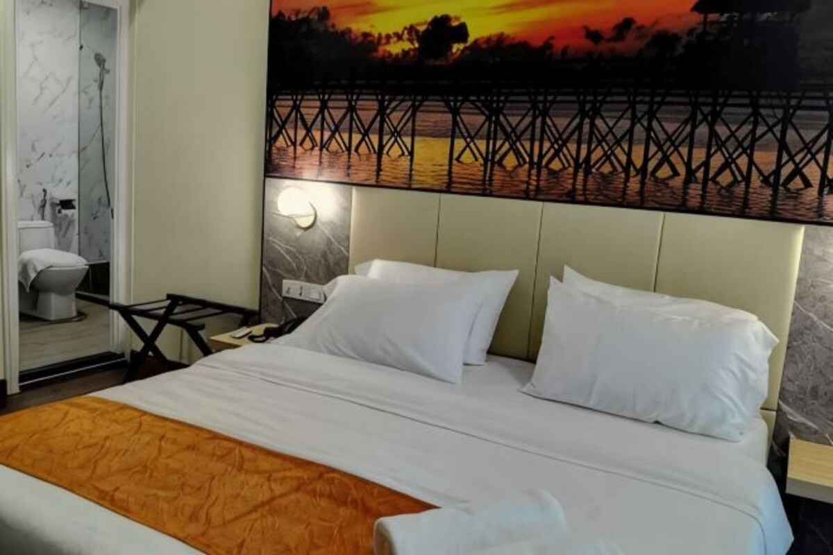 Ein Hotelzimmer mit Bett, Nachttisch, Wandlampe und einem Gemälde eines Sonnenuntergangs über einer Brücke.