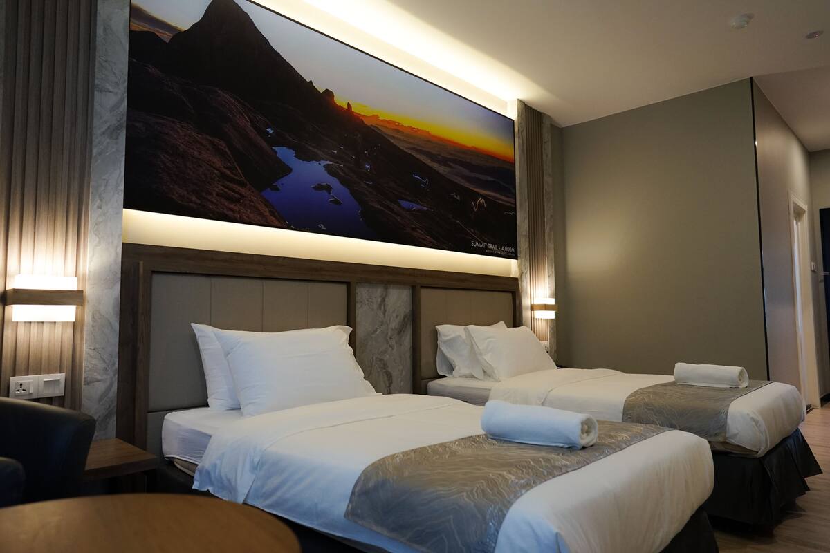 A hotel room with two beds, a large painting of a mountain landscape, and a wooden headboard.