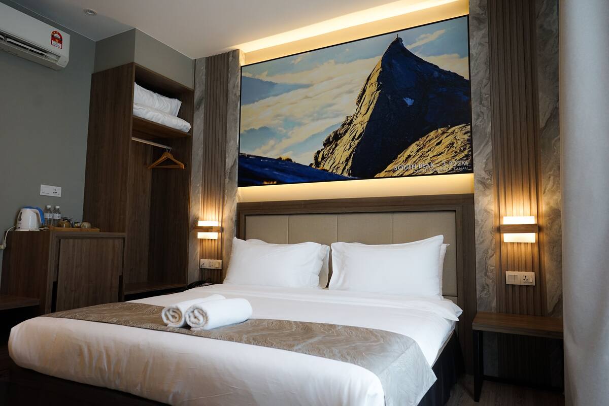 A hotel room with a large bed, a wooden wardrobe, a desk with a kettle, and a painting of a mountain.