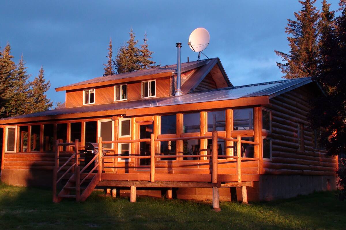 Uma cabana de toras com um deck de madeira, uma antena parabólica e uma escadaria que leva à entrada principal.