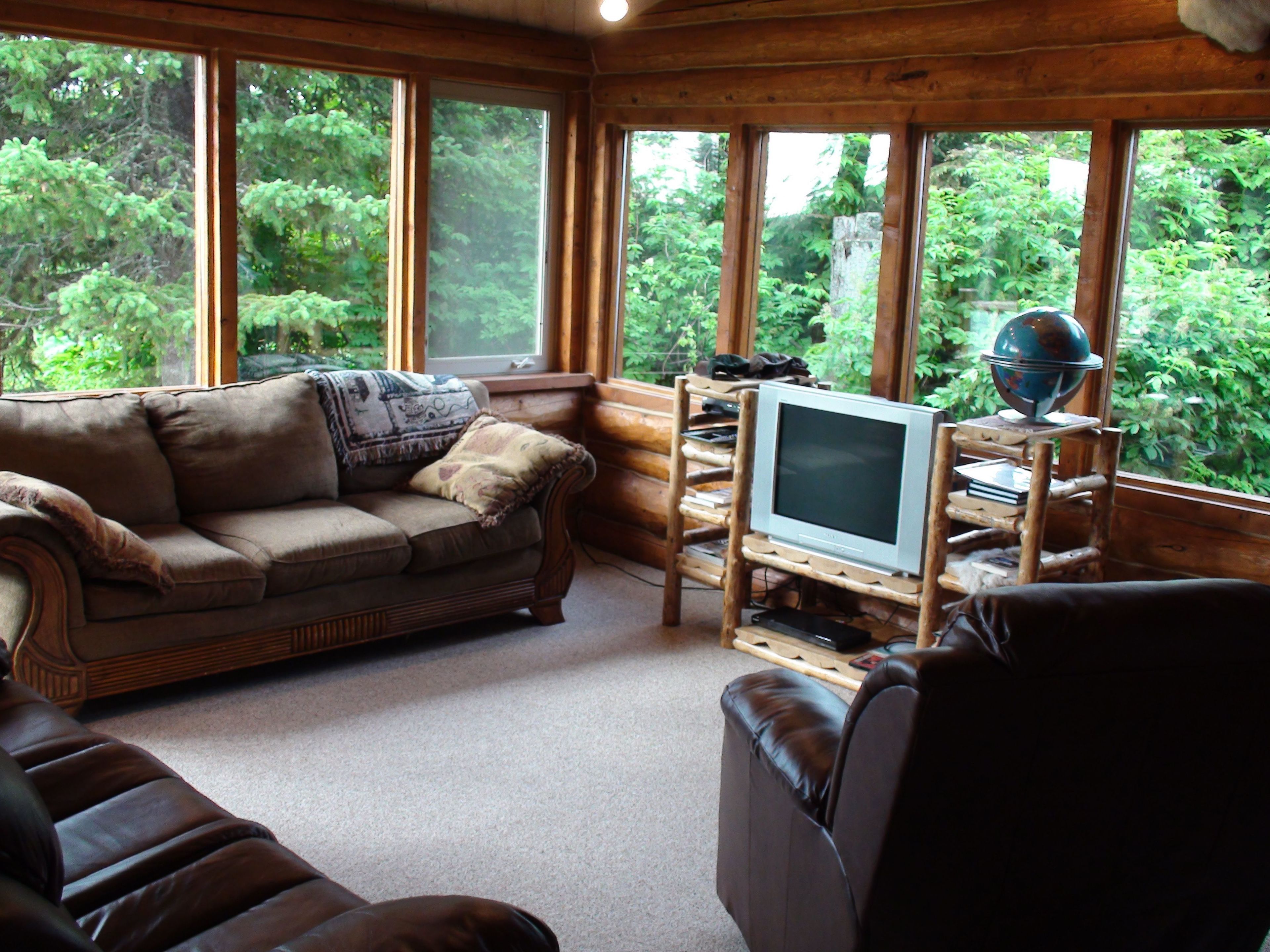 A cozy cabin interior with a brown leather sofa, a wooden coffee table, and a television on a wooden stand. Large windows offer a view of lush greenery outside.