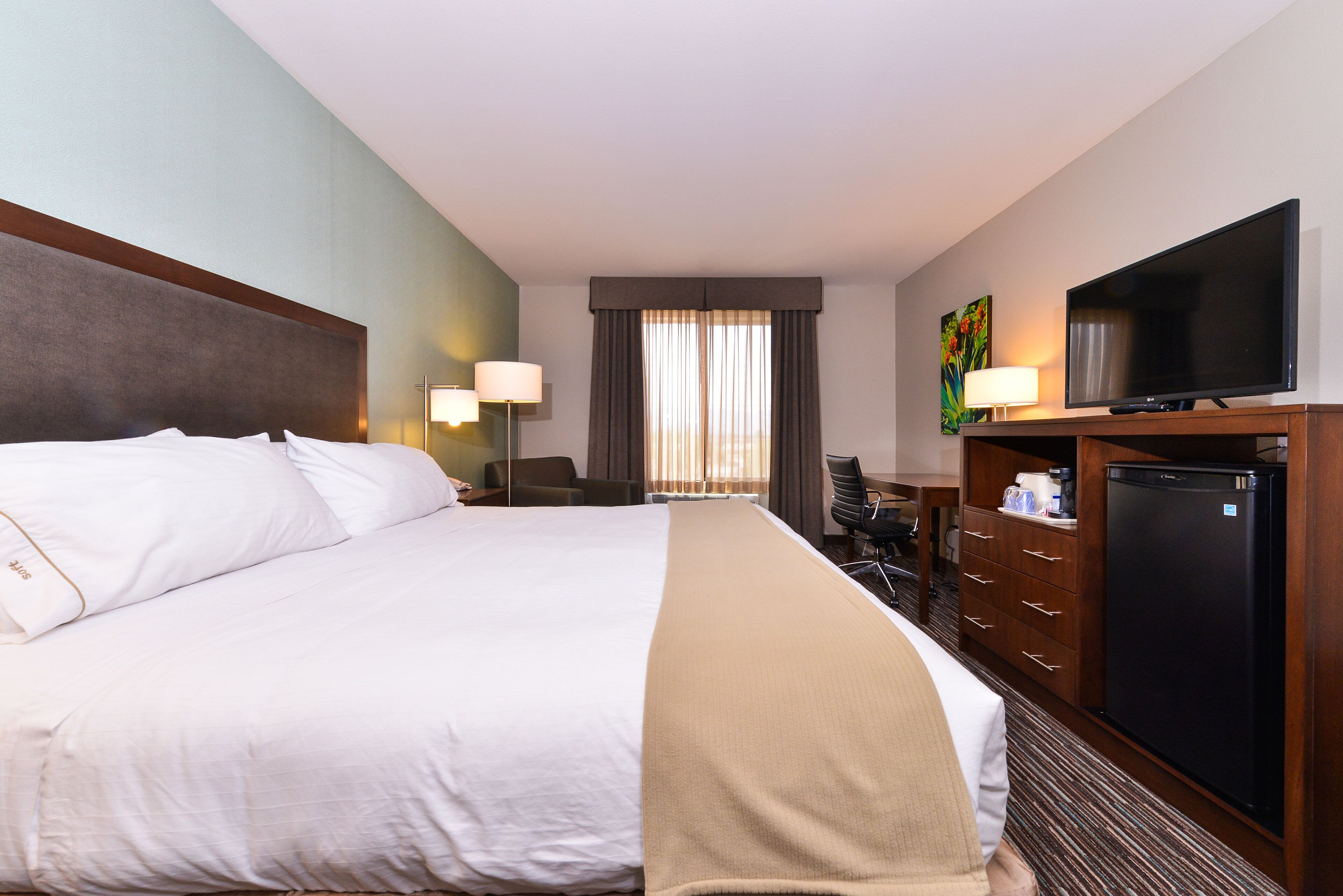 Quarto de hotel com uma cama grande, uma escrivaninha com computador, televisão e minibar.