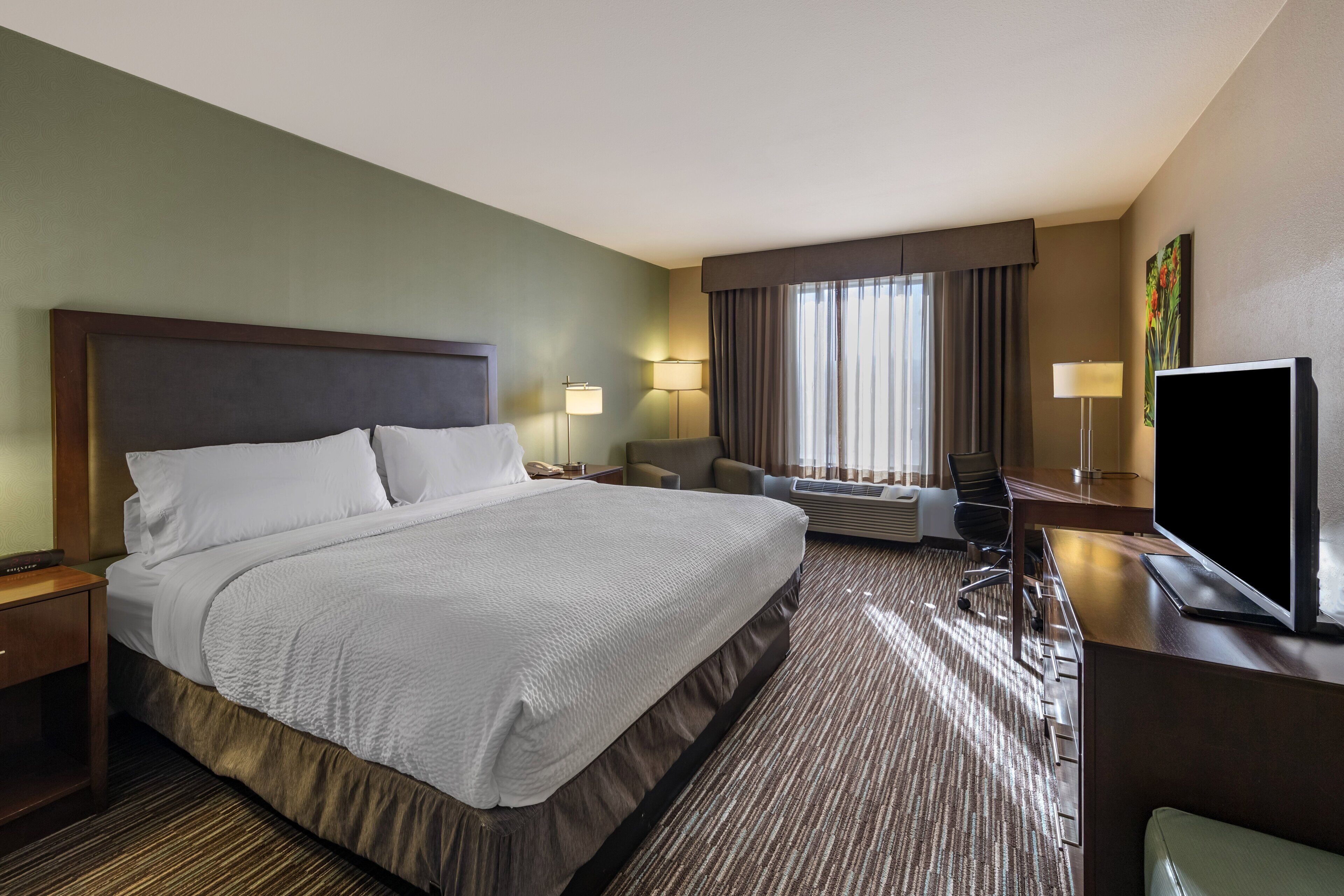 Ein Hotelzimmer mit einem großen Bett, einem Schreibtisch mit Fernseher, einem Sessel und einer Schreibtischlampe.