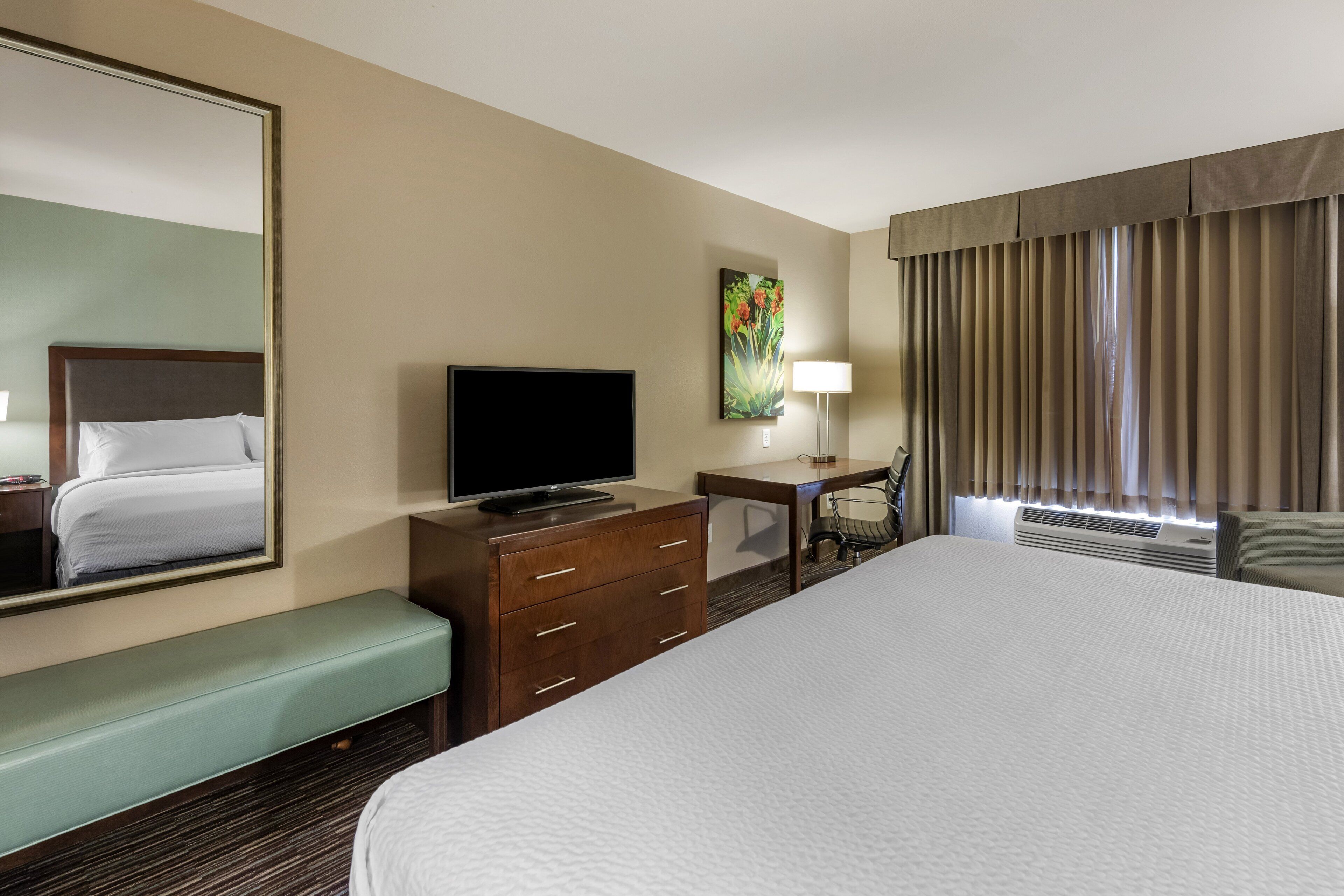 A hotel room with a large bed, a television, a desk with a chair, a lamp, and a large window with curtains.