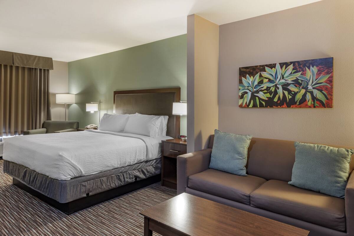 A hotel room with a bed, a sofa, a coffee table, a desk with a lamp, and a painting on the wall.
