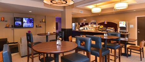 A hotel lobby with round tables and chairs, a bar area, and a television displaying capsules.