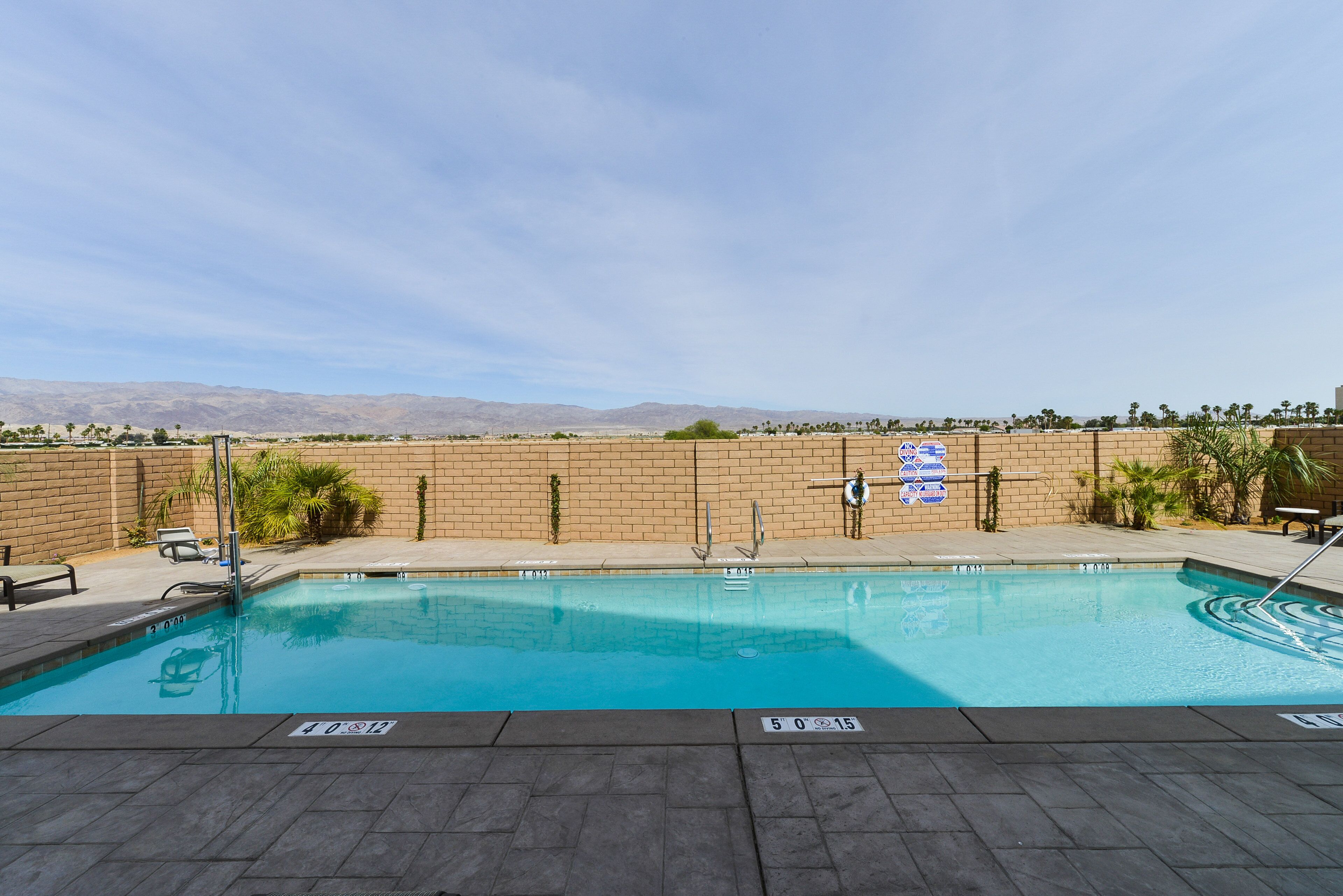 Una piscina con agua cristalina, rodeada por un muro de ladrillo y palmeras, con montañas al fondo.