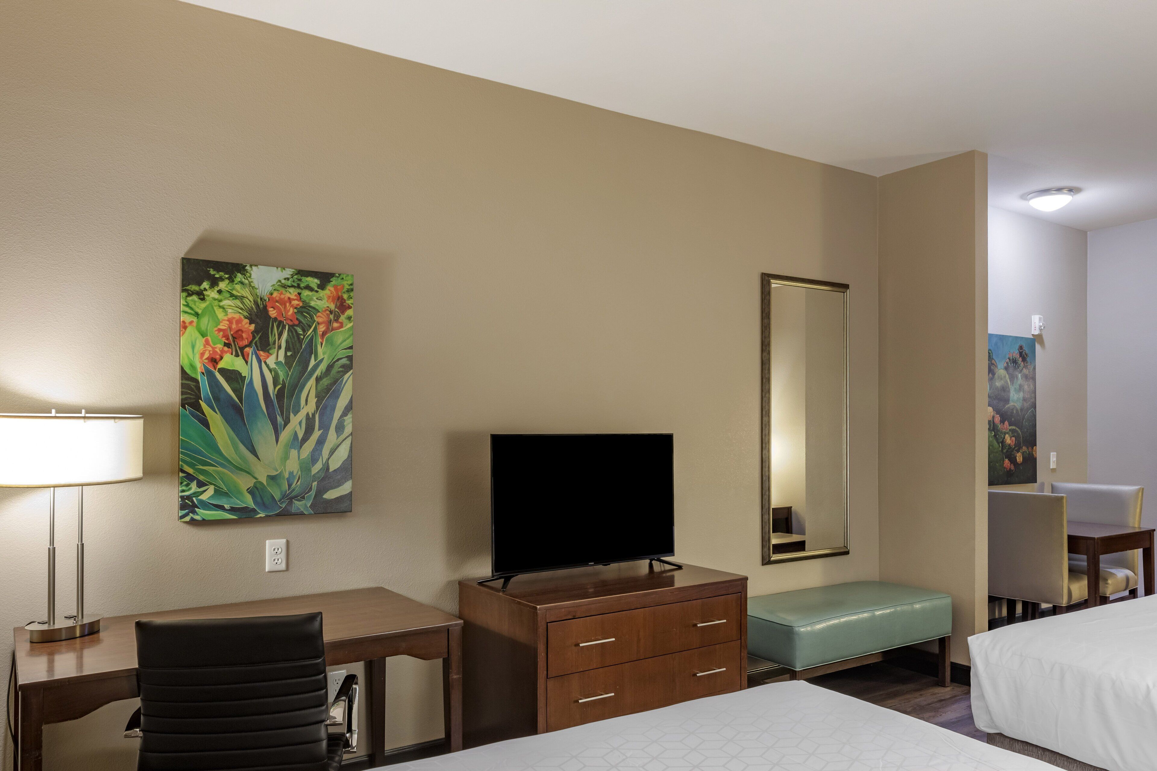 A hotel room with a bed, a desk with a chair, a television, a dresser, a bench, and a large mirror.