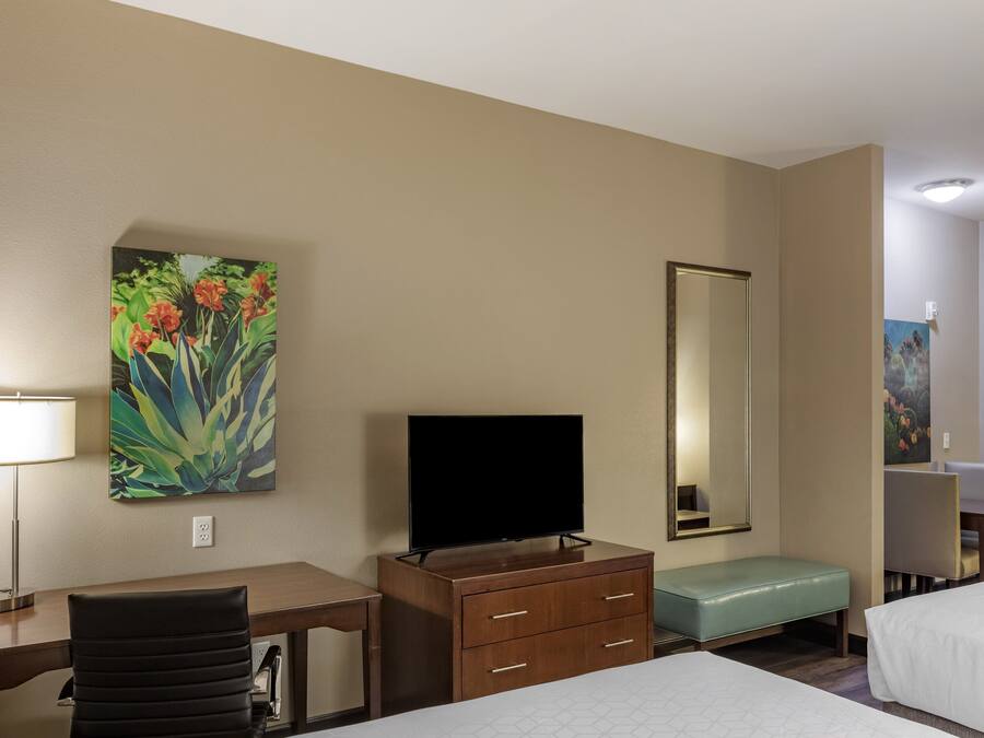 A hotel room with a bed, a desk with a chair, a television, a dresser, a bench, and a large mirror.