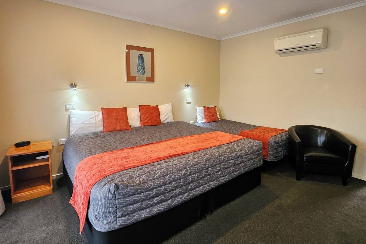 Quarto de hotel com uma cama grande, duas mesas de cabeceira, uma cadeira, um aparelho de ar condicionado e um quadro na parede.