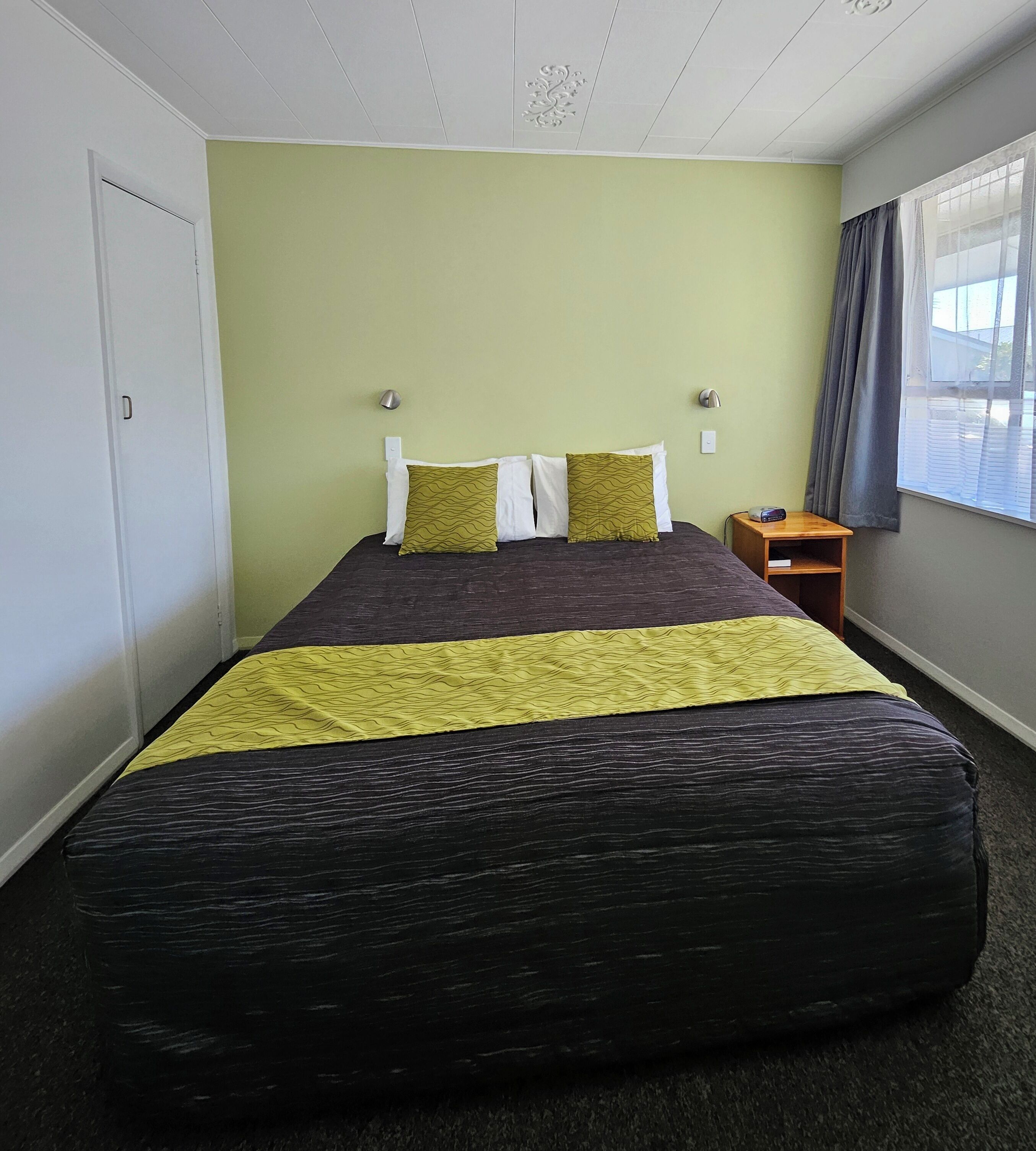 A hotel room with a bed, a nightstand, a lamp, a window with curtains, and a door.