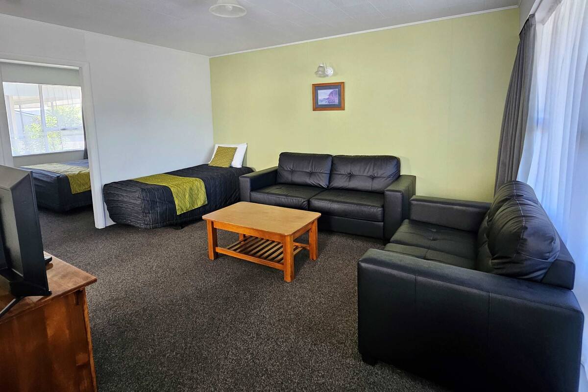 Uma sala de estar mobiliada, com um sofá, um pufe, uma mesa de centro de madeira e uma televisão sobre um suporte.