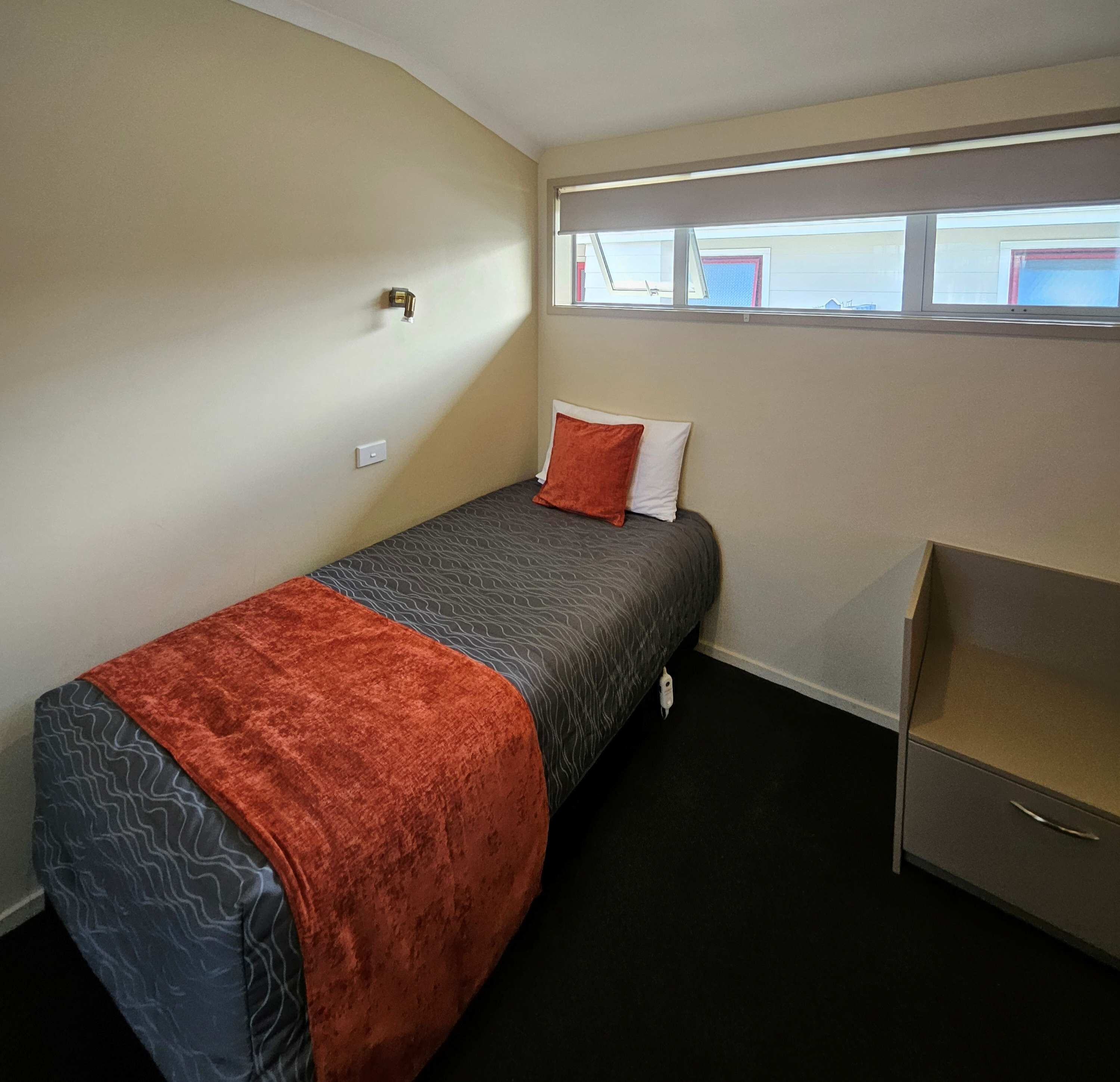 Una cama individual con una colcha estampada, una manta roja y una almohada. Hay un escritorio pequeño con cajones y una ventana sobre la cama.