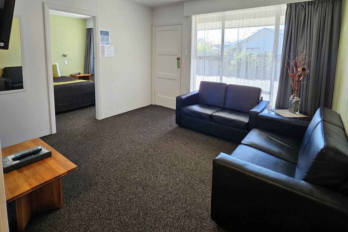 Um quarto com carpete escuro, sofá preto, mesa de madeira com um controle remoto e janela com cortinas.