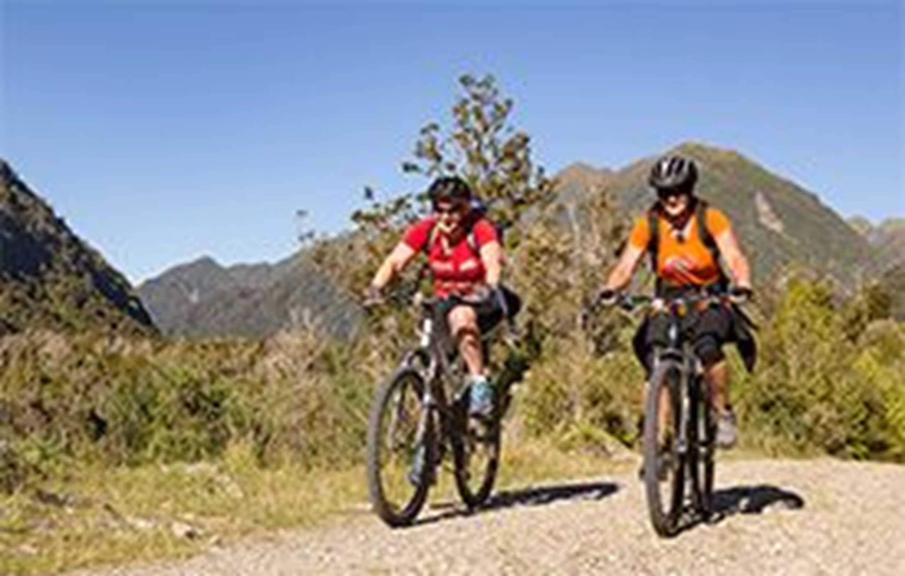 Two cyclists on a dirt path with mountains in the background.