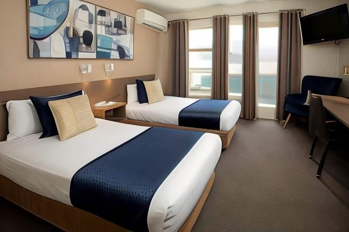 Habitación de hotel con dos camas, un escritorio, una silla, un televisor y un ventanal con cortinas.