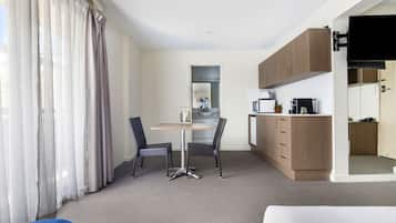 A compact hotel room with a kitchenette, a dining area with two chairs, a sofa, and a large window with sheer curtains.
