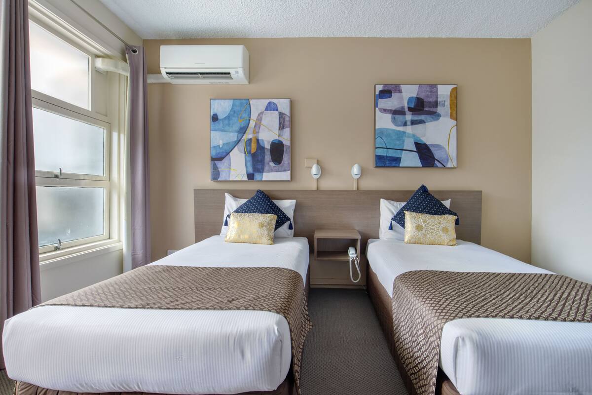 Quarto de hotel com duas camas, uma janela com cortinas, um aparelho de ar condicionado e obras de arte abstratas nas paredes.