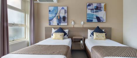 A hotel room with two beds, a window with curtains, an air conditioning unit, and abstract artwork on the walls.