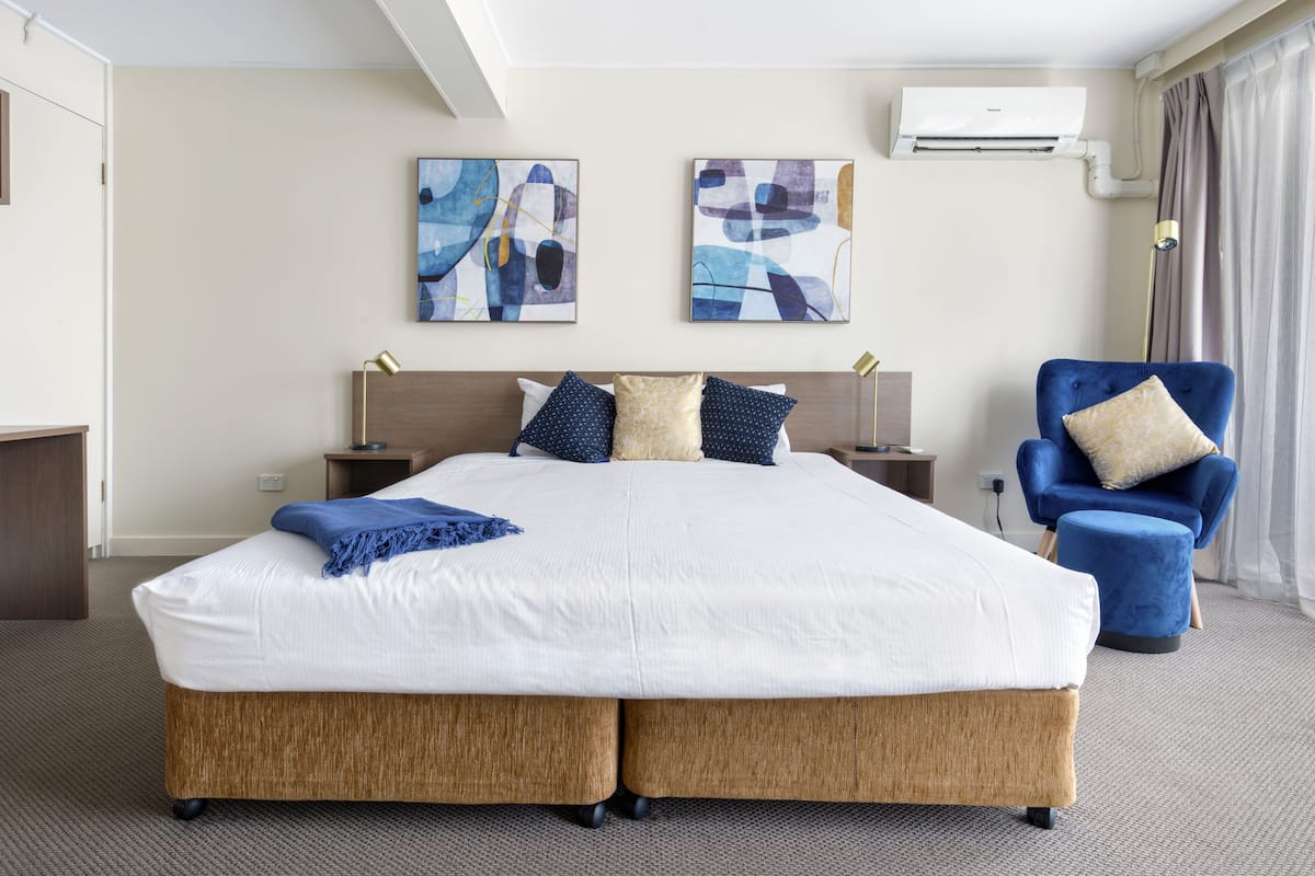 Quarto de hotel com uma cama grande, duas mesas de cabeceira, uma poltrona azul e obras de arte abstratas nas paredes.