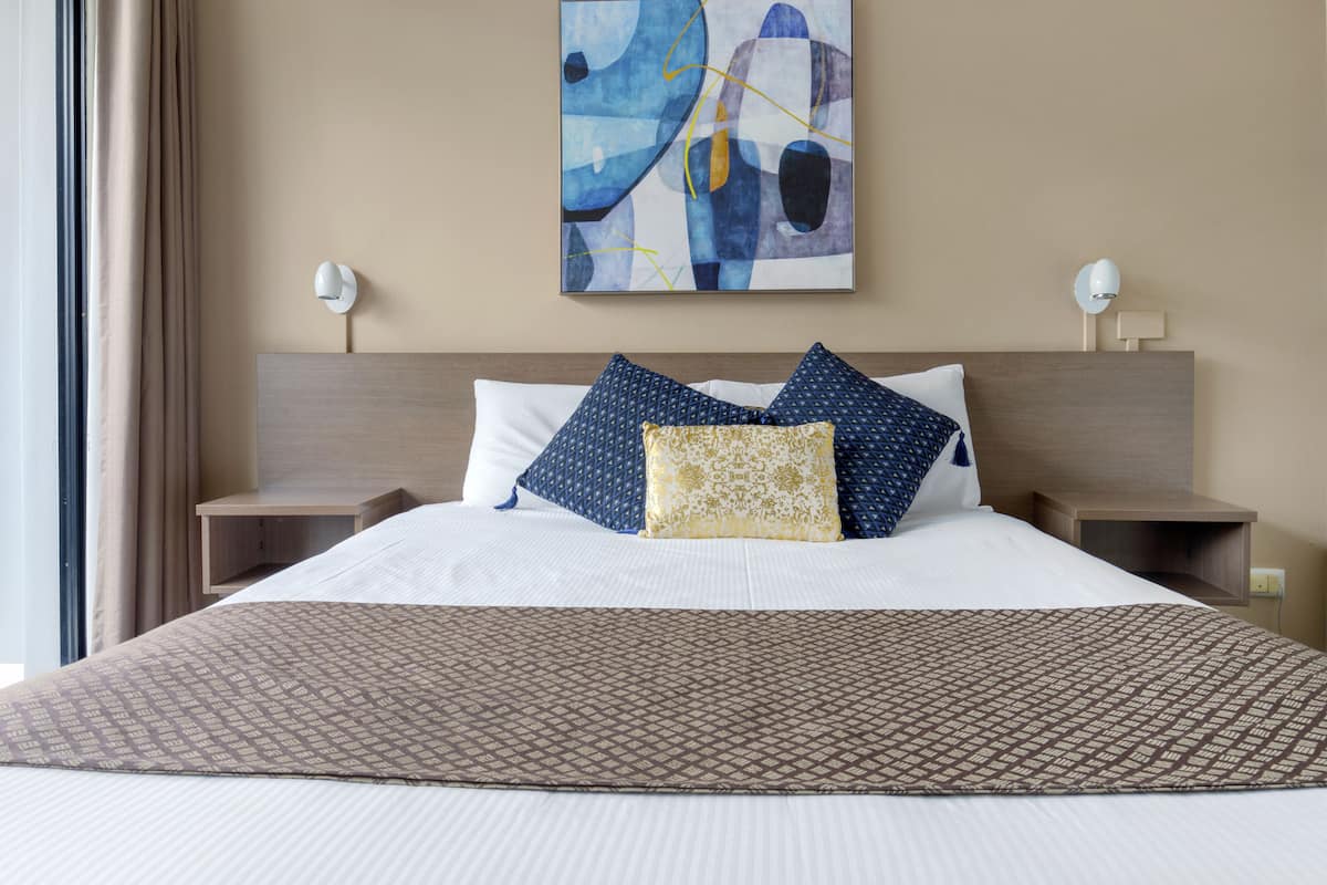 Quarto de hotel com uma cama grande, duas mesas de cabeceira, um quadro abstrato pendurado na parede e uma janela com cortinas.