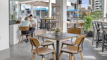 An outdoor cafe with wooden tables and chairs, potted plants, and a view of the cityscape.