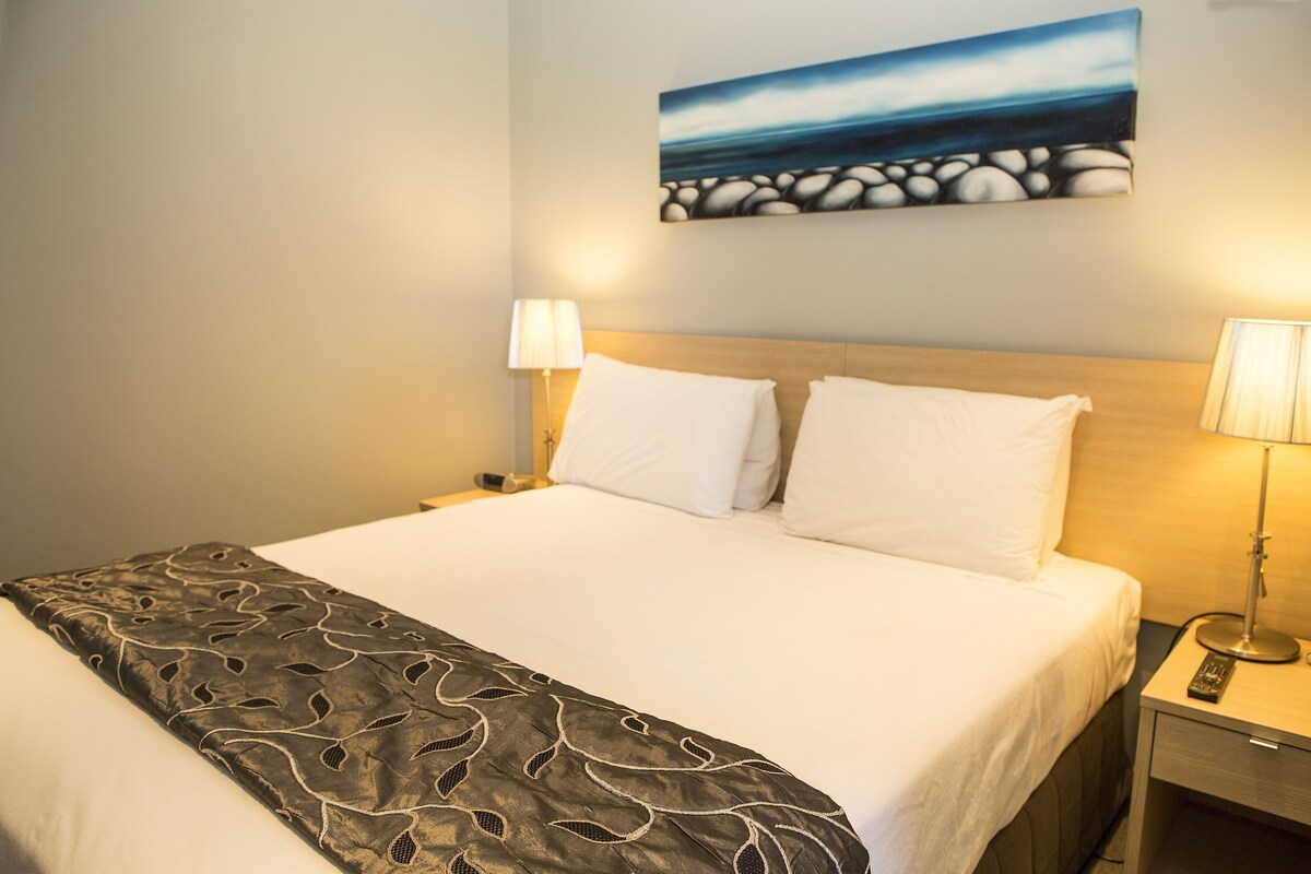 A hotel room with a bed, two bedside lamps, a painting on the wall, and a remote control on the nightstand.