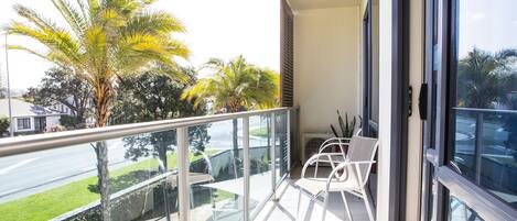 A balcony with a glass railing, white chairs, and a view of a street with palm trees.