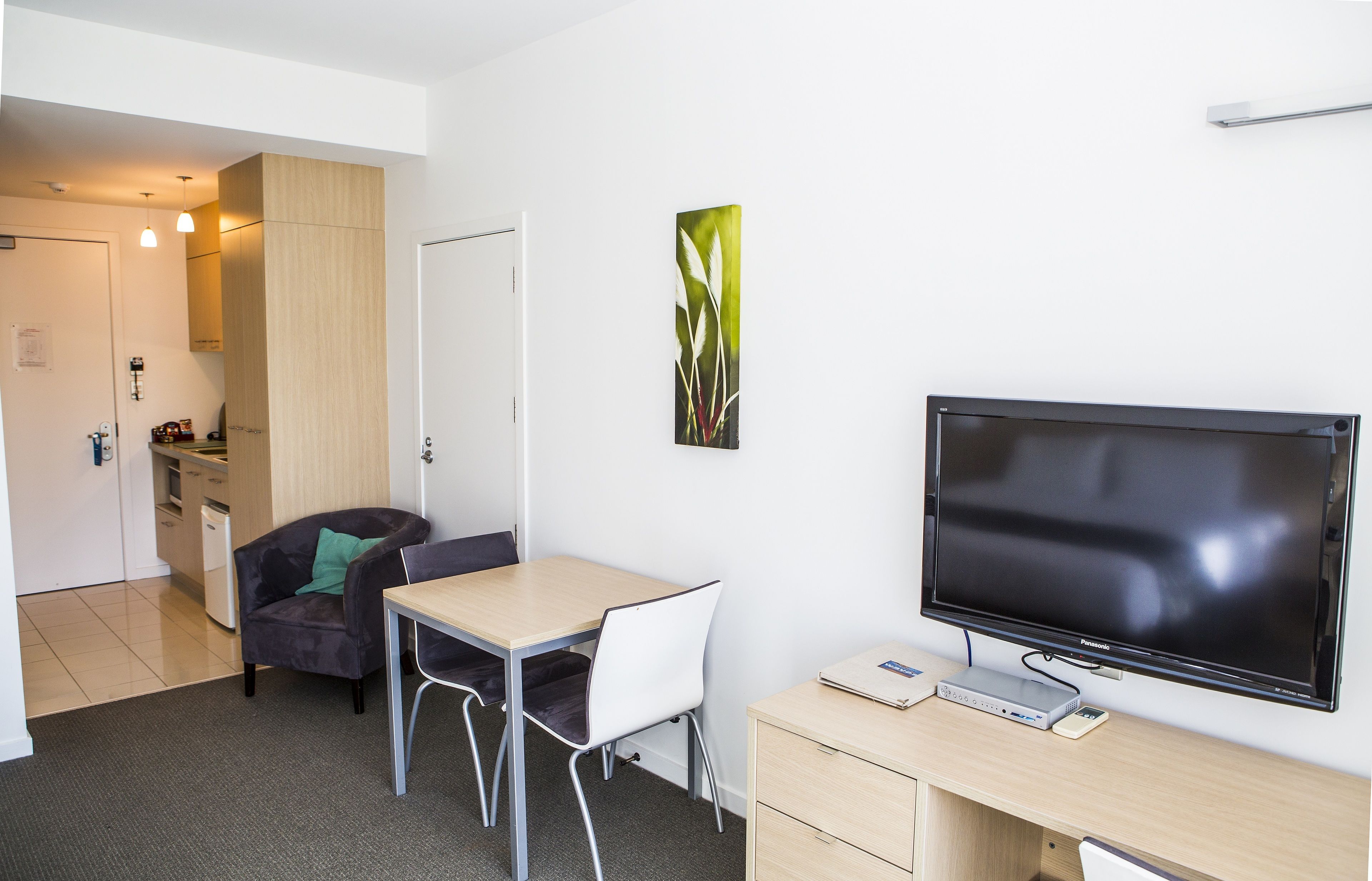 A modern room with a flat-screen TV, a desk, two chairs, a small cabinet, and a door leading to a kitchenette.