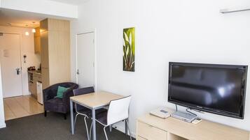A modern room with a flat-screen TV, a desk, two chairs, a small cabinet, and a door leading to a kitchenette.