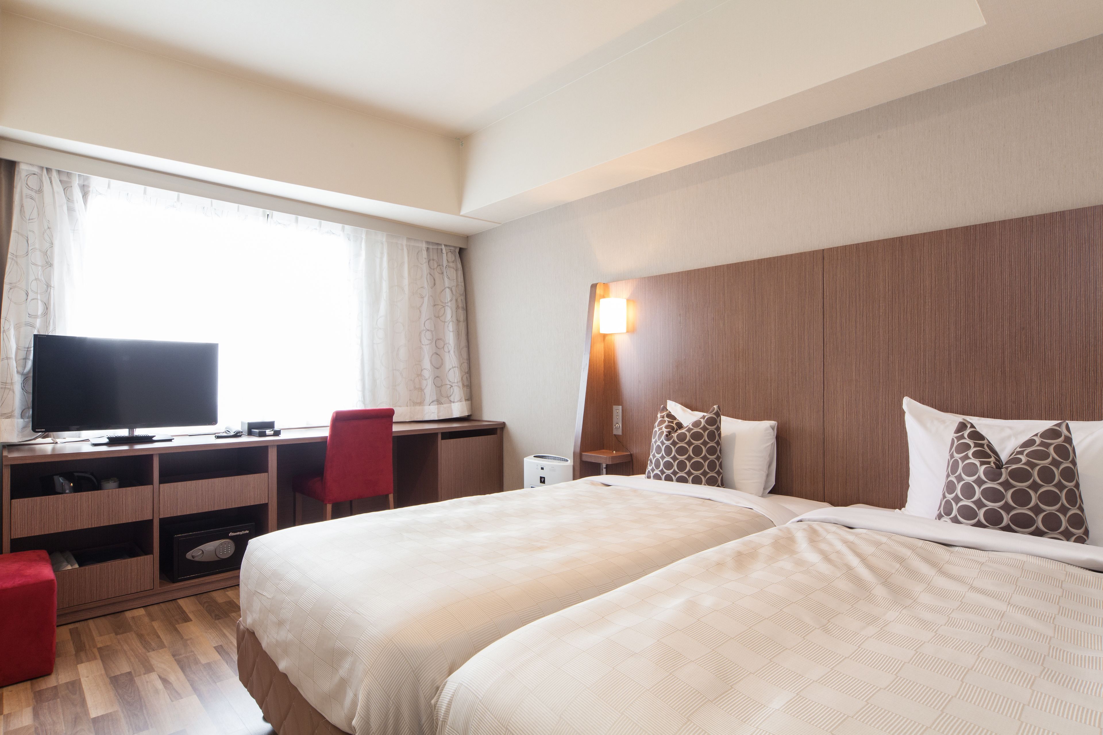 A hotel room with a bed, a desk with a television, a chair, and a bedside table.